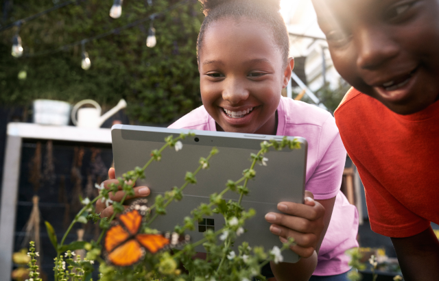 Vrouwelijke K-12 student in een tuin met een Surface Go tijdens het maken van een foto van een vlinder. Lachende mannelijke K-12 student staat naast haar.